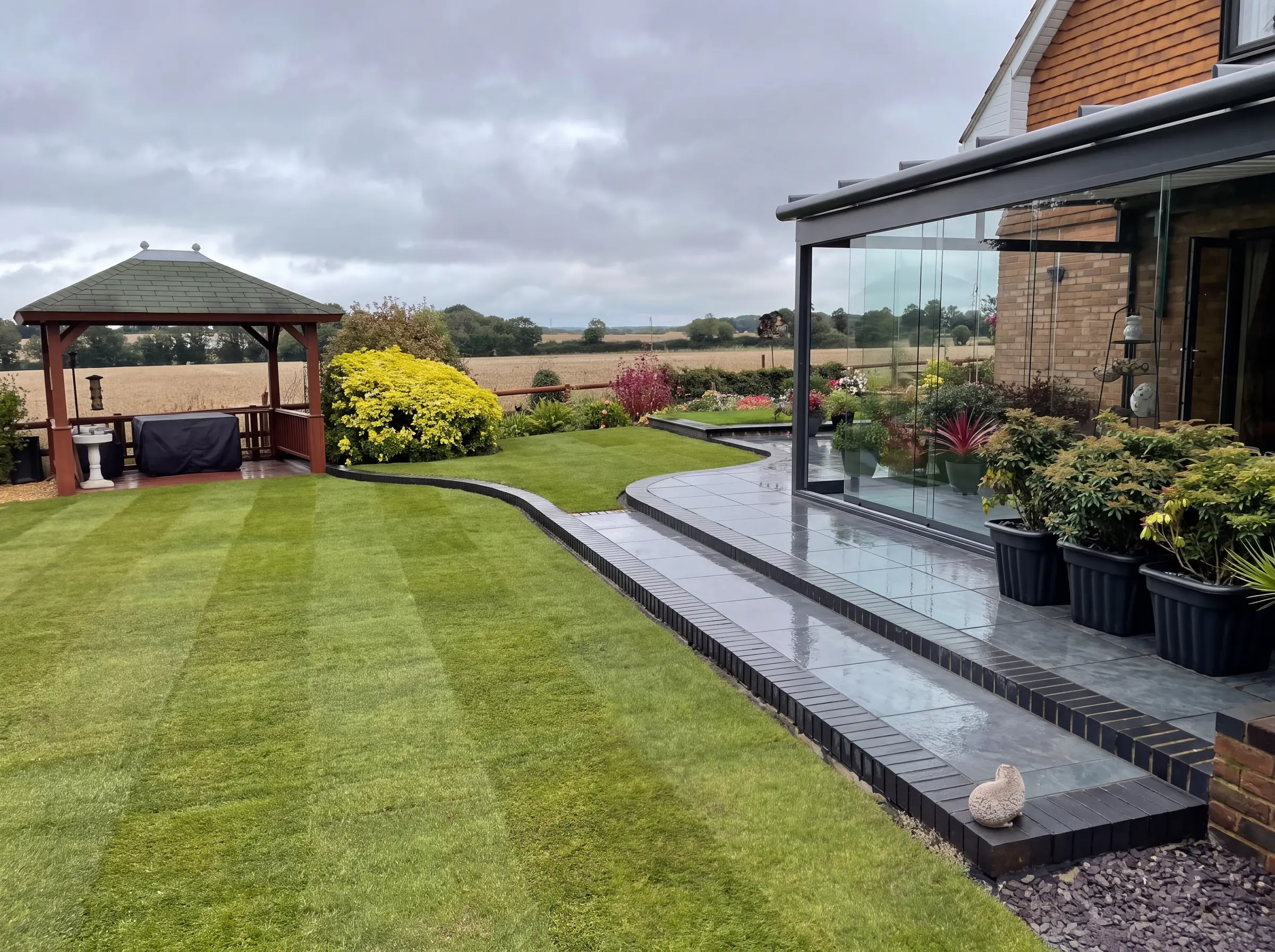 Striped lawn and porcelain terrace beside a glass garden room, with a pavilion beyond