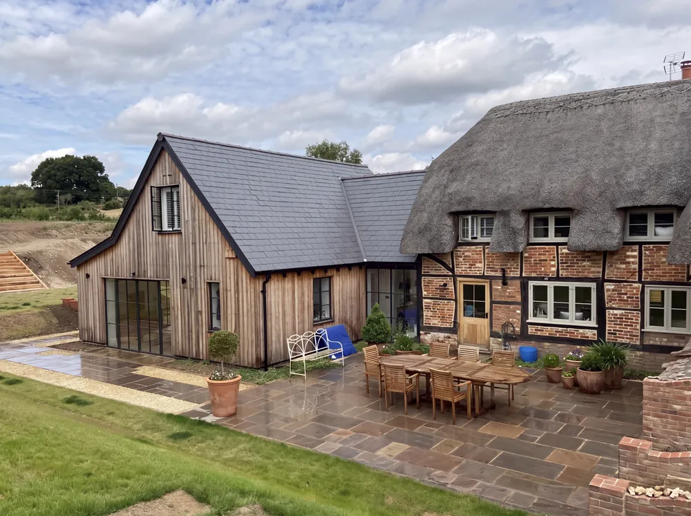 New timber home beside a thatched cottage, with a paved terrace and lawn